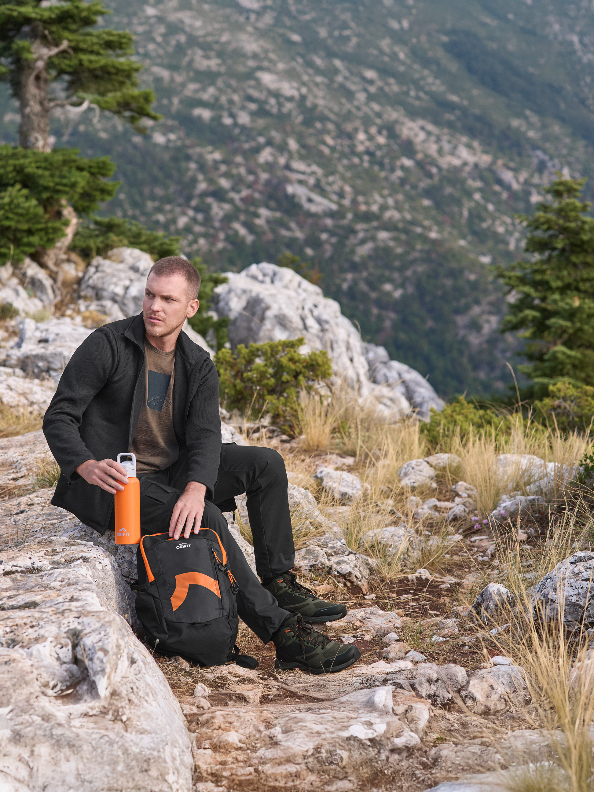 Homme en veste et pantalon d'extérieur Crivit, tenant une gourde orange Crivit, avec un sac à dos Crivit.
