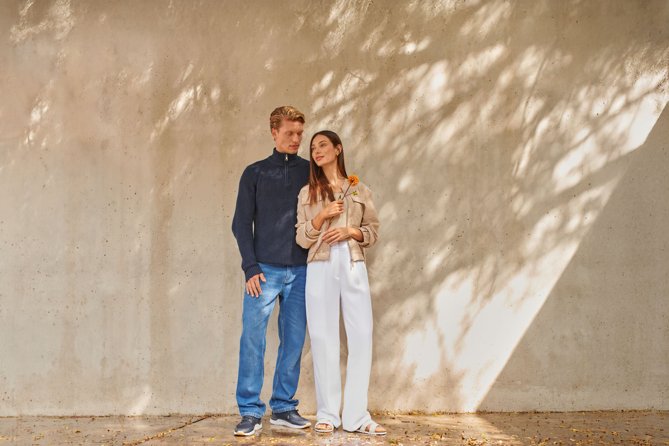 Koppel in casual kleding: man in blauwe trui en jeans, vrouw in beige jasje en witte broek.