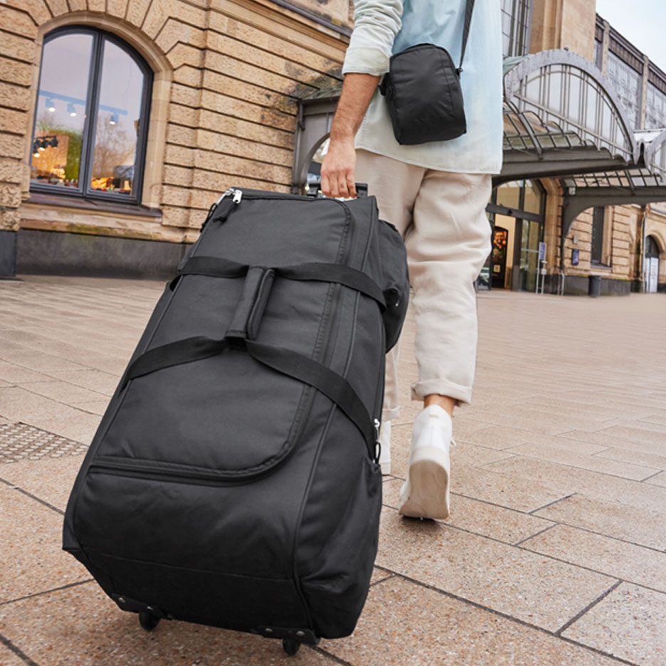 Een reiziger trekt een grote zwarte koffer op wielen voor een treinstation.