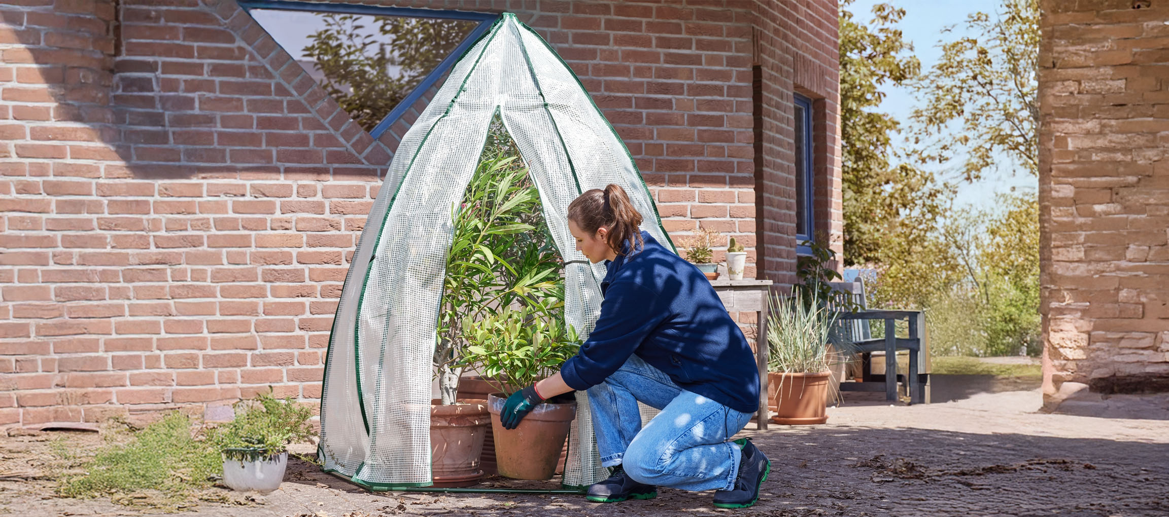 Vrouw plaatst potplanten in een kas bij een bakstenen gebouw.