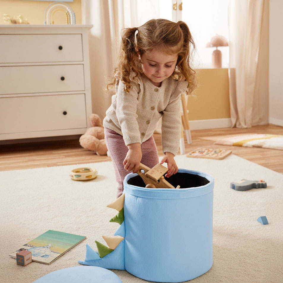 Une enfant range des jouets en bois dans un bac de rangement bleu en forme de dinosaure dans une chambre.