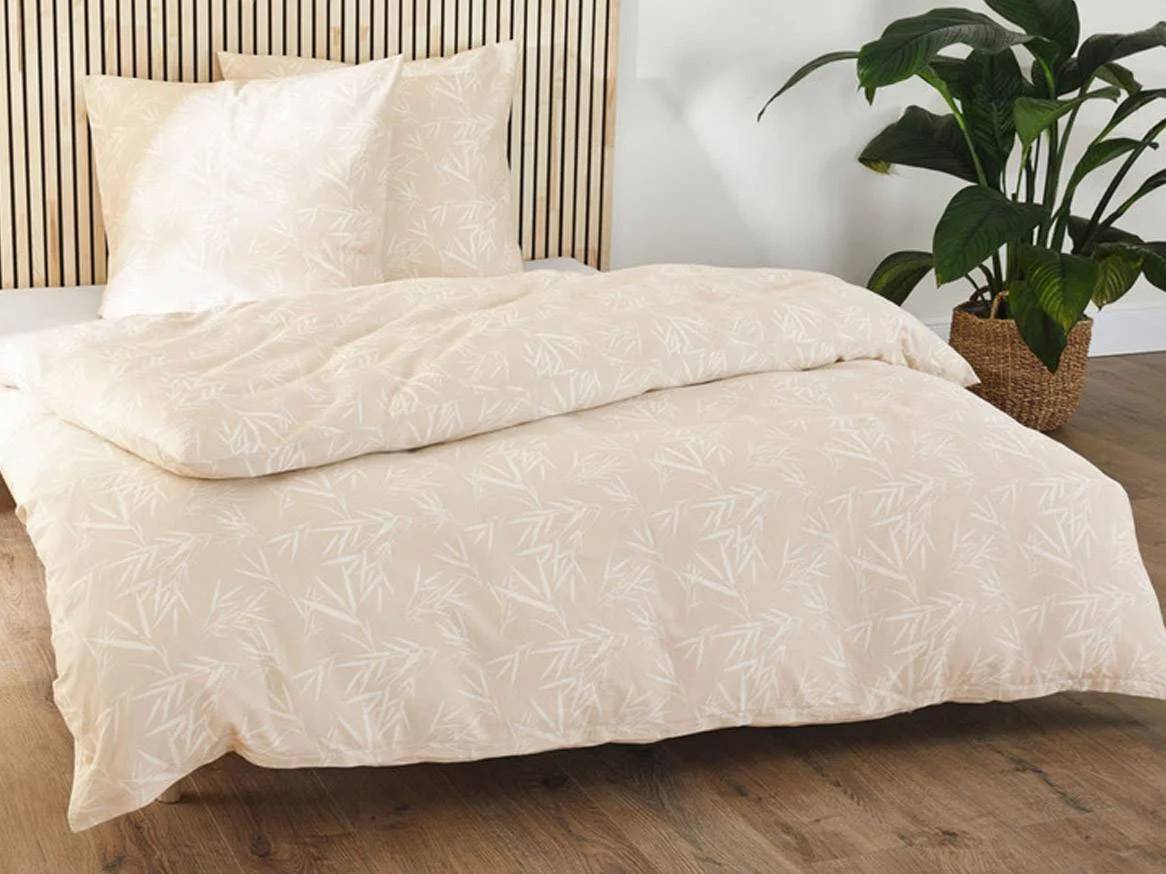 Crèmekleurig beddengoed met bamboepatroon op een bed met houten hoofdeinde en plant.