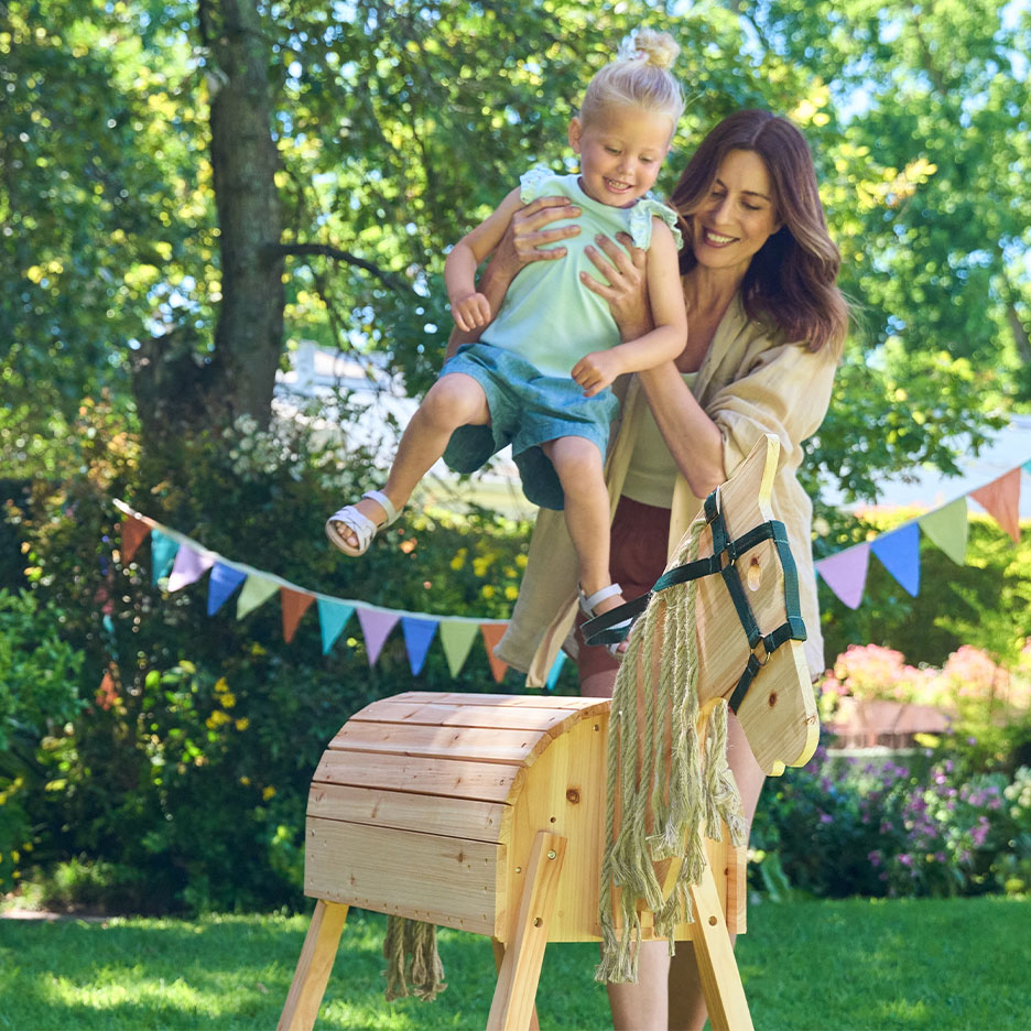 Vrouw en meisje spelen met een houten hobbelpaard in een tuin met vlaggetjes.