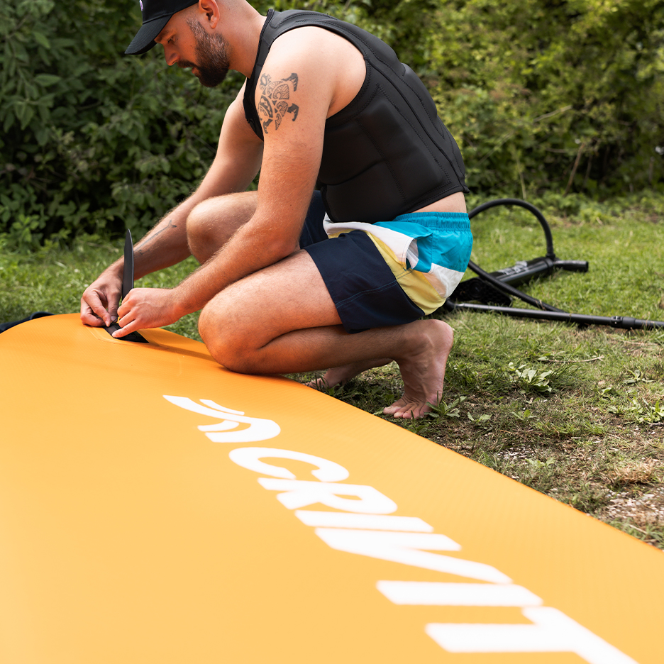 Man monteert een oranje paddleboard, draagt een zwemvest en zwemshorts.
