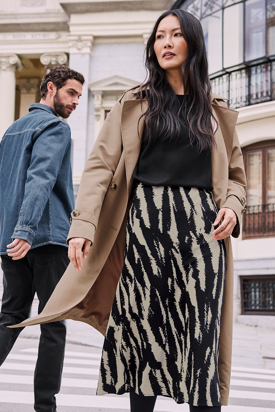 Een vrouw in een beige trenchcoat en zebraprint rok, met een man in een spijkerjack achter haar.