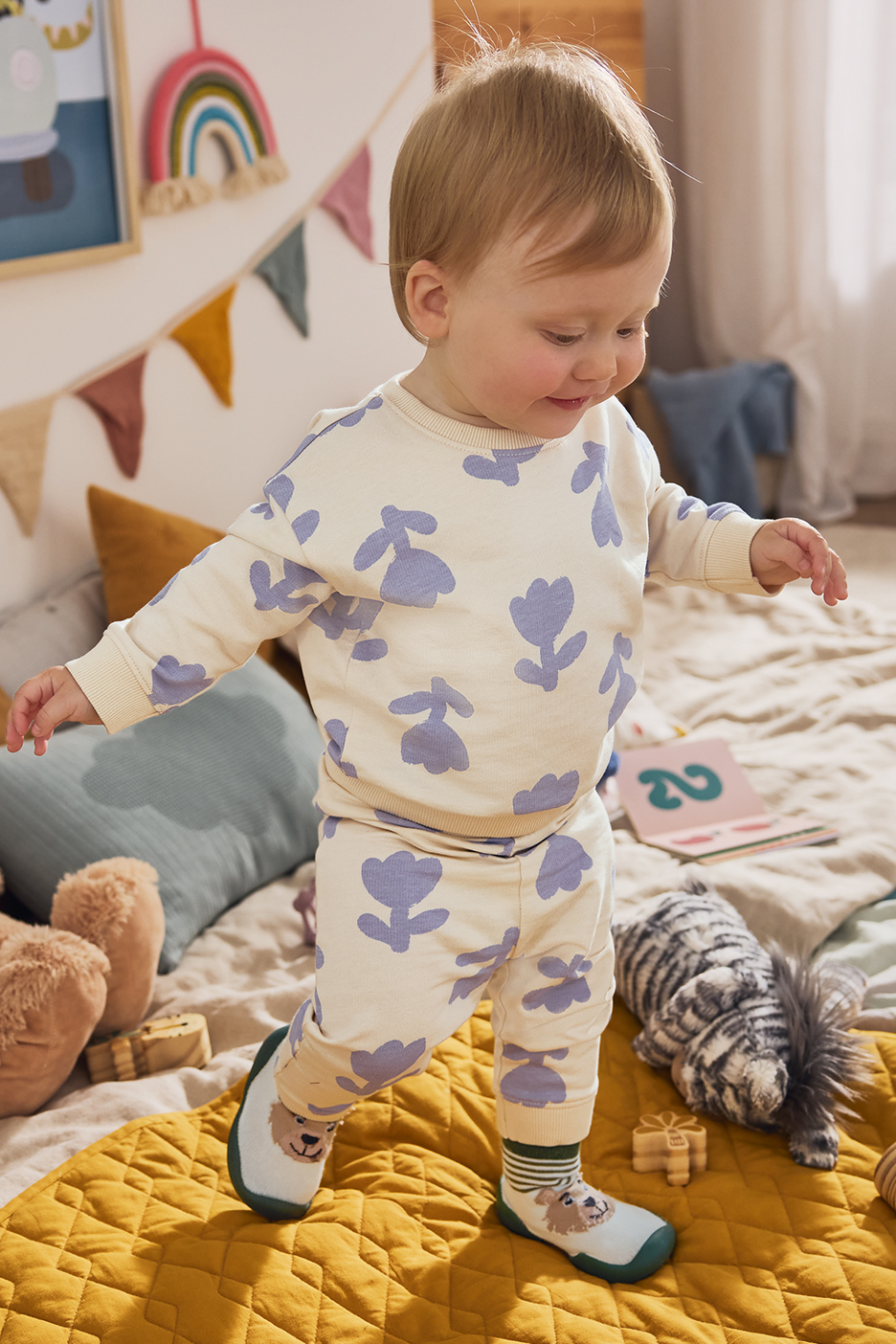Een baby in een crèmekleurige pyjama met paarse bloemen, staand op een gele deken.