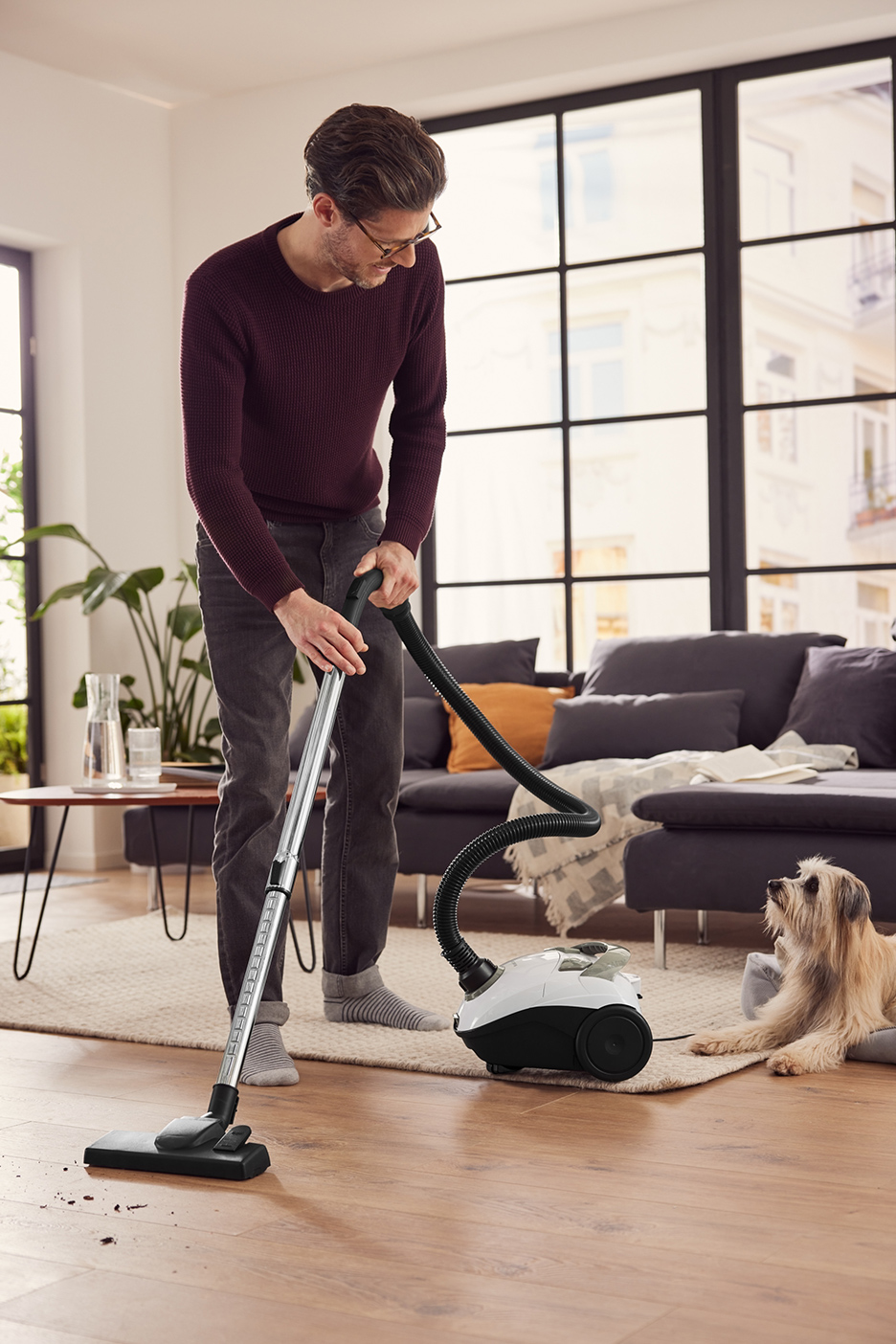 Een man stofzuigt een houten vloer, met een hond die in de buurt ligt.