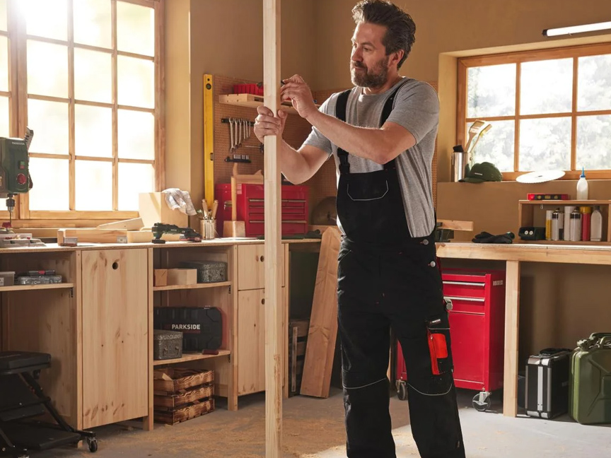 Un homme en salopette de travail dans un atelier de menuiserie avec des outils et du bois.