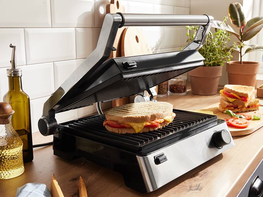 Een open paninigrill met een kaas-tomaatsandwich, en een andere gesneden sandwich.