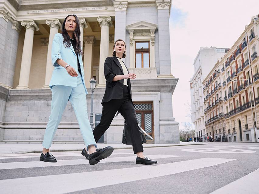 Twee vrouwen in pakken en loafers steken een straat over voor een klassiek gebouw.