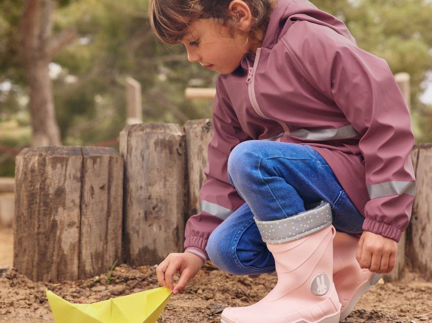 Kind in roze regenjas en roze laarzen speelt met een papieren bootje.