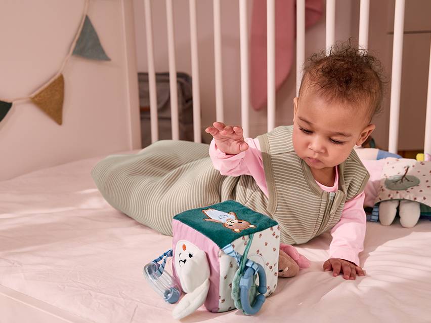 Een baby in een gestreepte slaapzak speelt met een activiteitenkubus in een wieg.