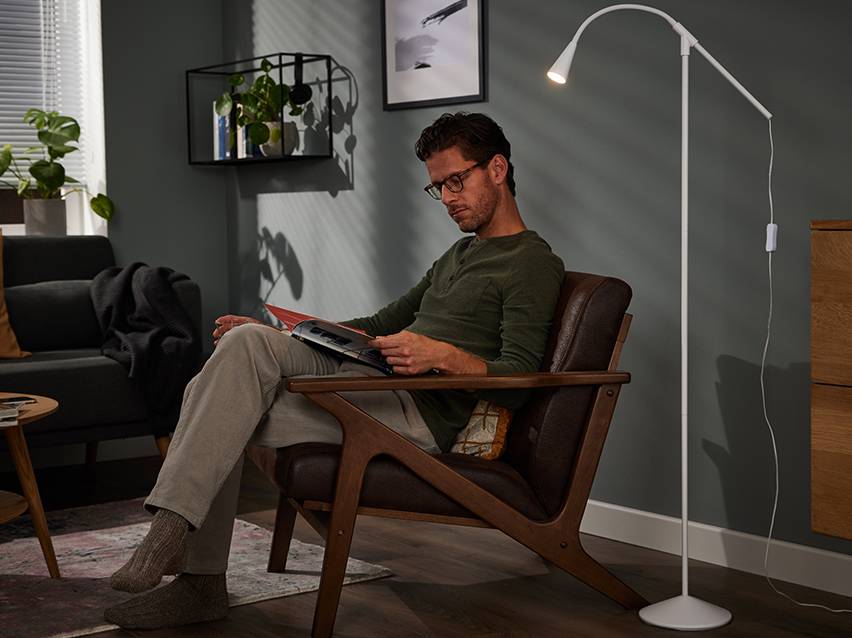 Een man leest in een fauteuil, verlicht door een witte vloerlamp in een woonkamer.