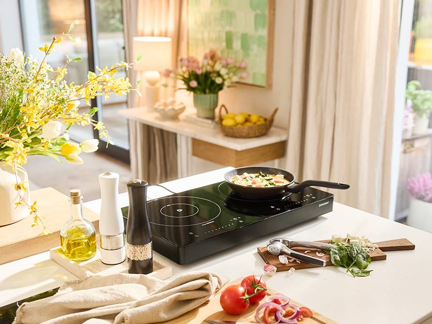 Table de cuisson à induction portable avec une poêle, des épices et des légumes frais sur un comptoir de cuisine.
