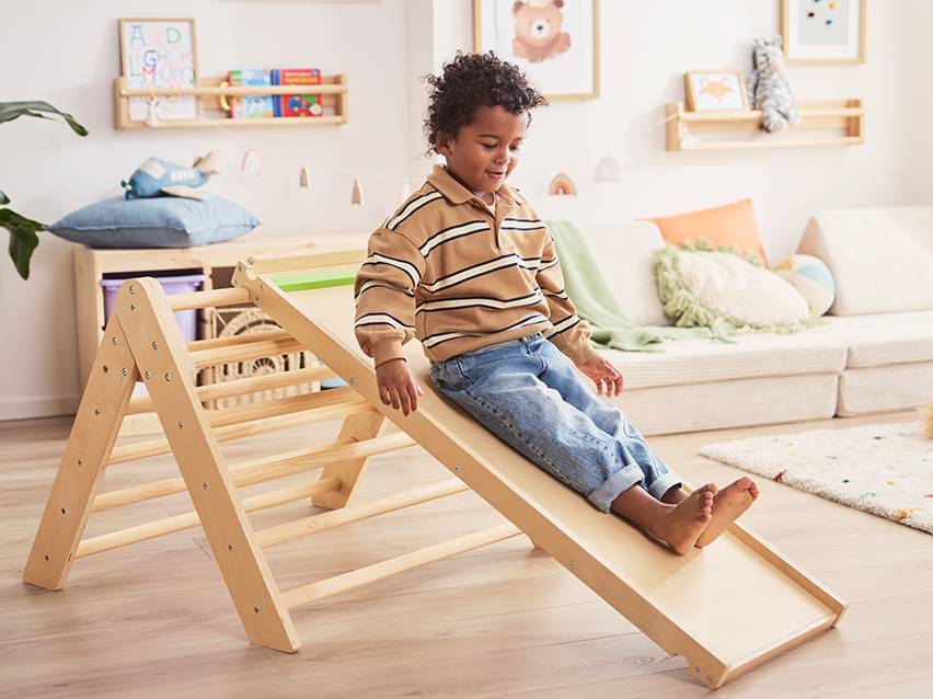 Een lachend kind glijdt van een houten speeltoestel in een kinderkamer met een bank en planken.