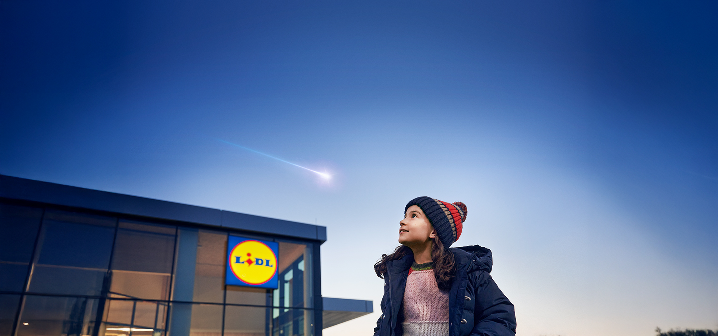 Meisje kijkt naar vallende ster boven Lidl-winkel, draagt wintermuts en jas.