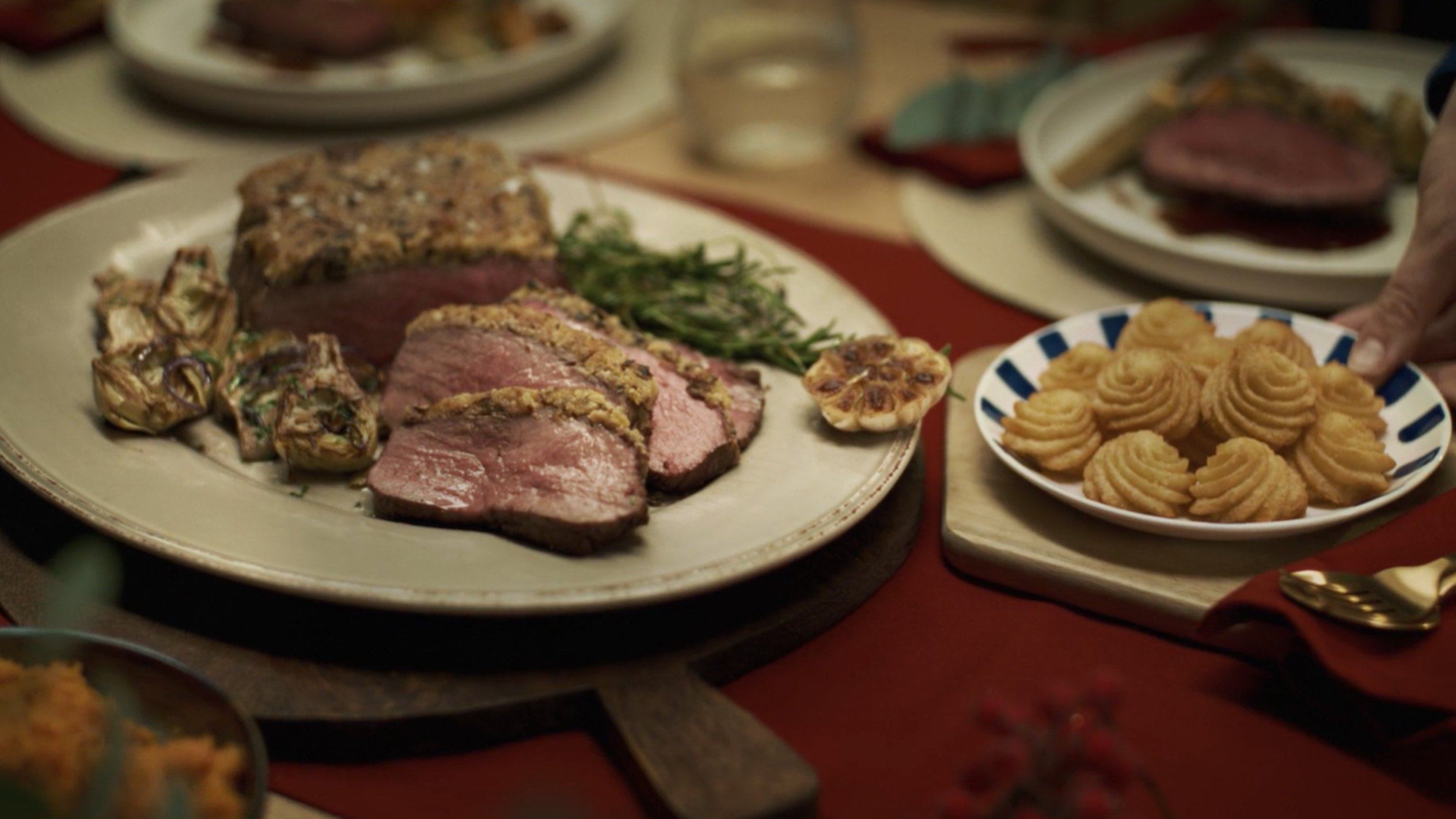 Feestelijk diner met rosbief, artisjokken en aardappelrozetten op een rode tafel.