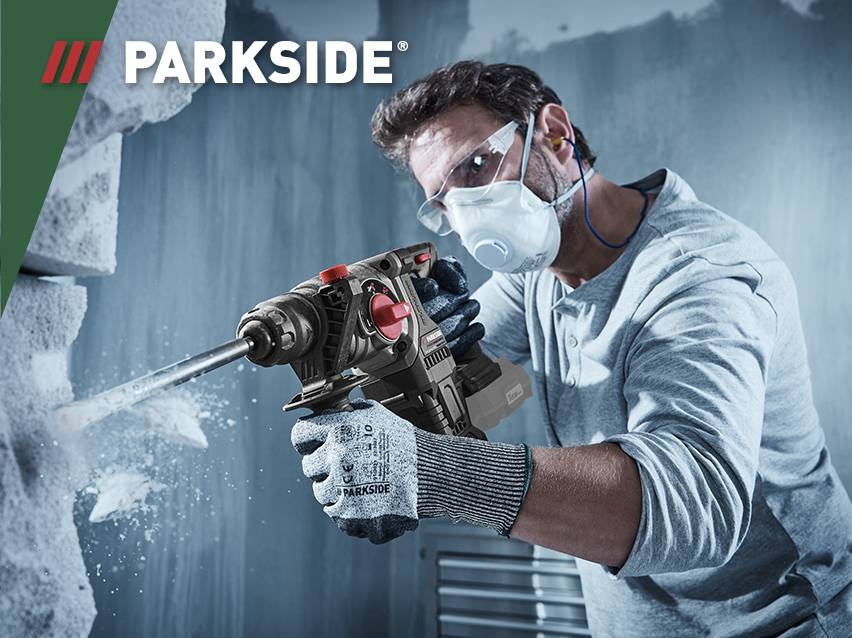 Een man met veiligheidsuitrusting gebruikt een Parkside boorhamer om in een muur te boren.