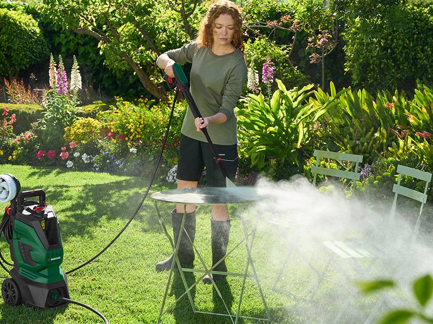 Vrouw reinigt tuintafel met hogedrukreiniger in zonnige tuin.