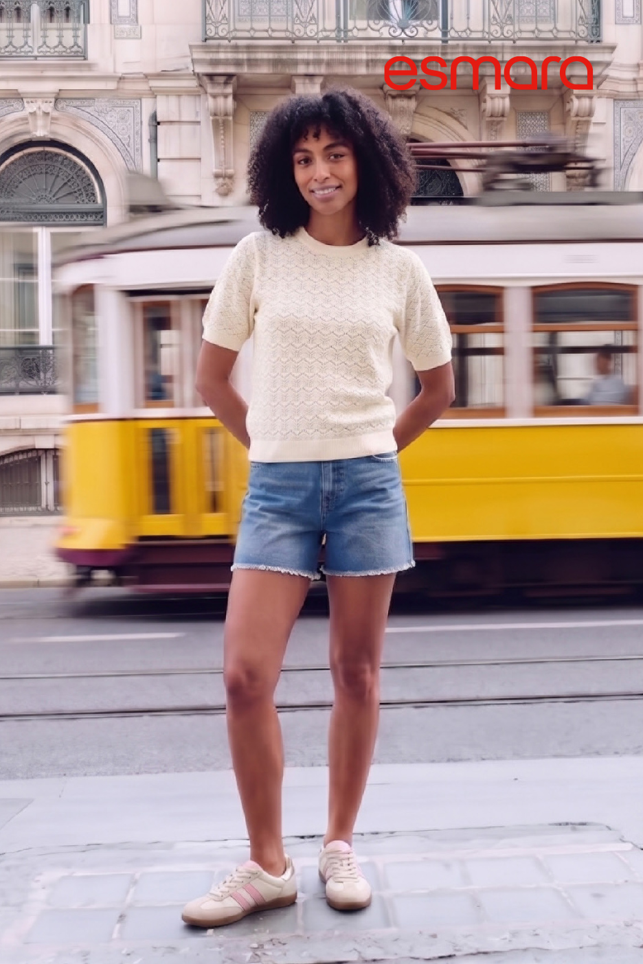 Vrouw in gebreide trui en korte broek, glimlachend voor een rijdende tram.