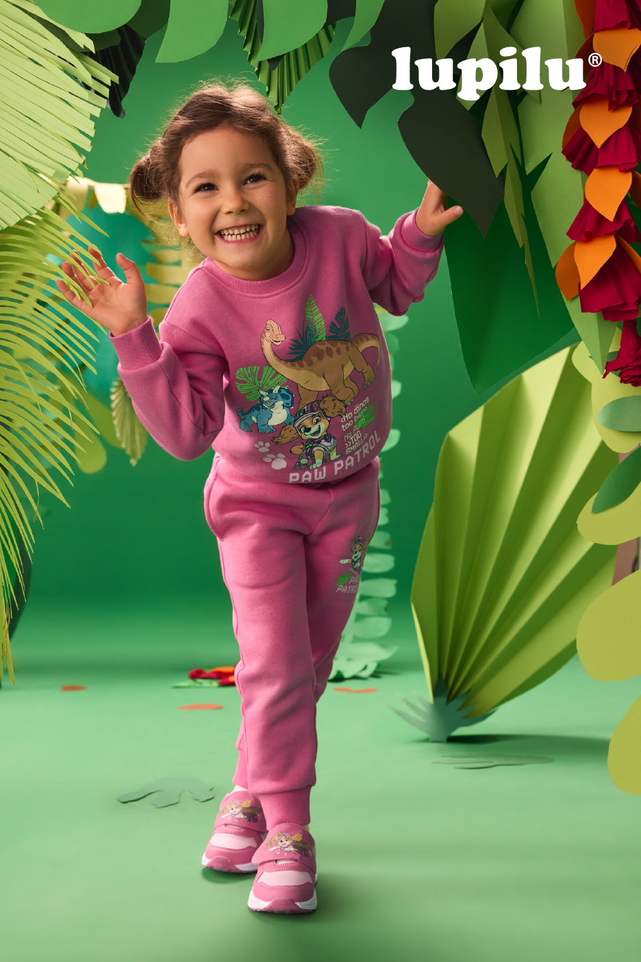 Lachend meisje in roze joggingpak met dinosaurus- en hondenprint, in een jungle-achtige setting.