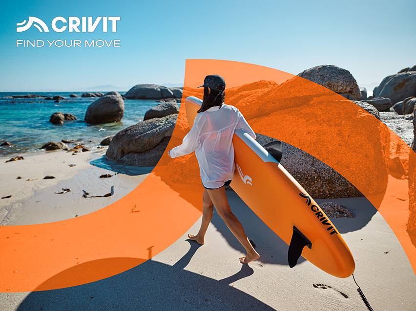 Vrouw loopt op zandstrand met oranje stand-up paddleboard, rotsen en zee op de achtergrond.