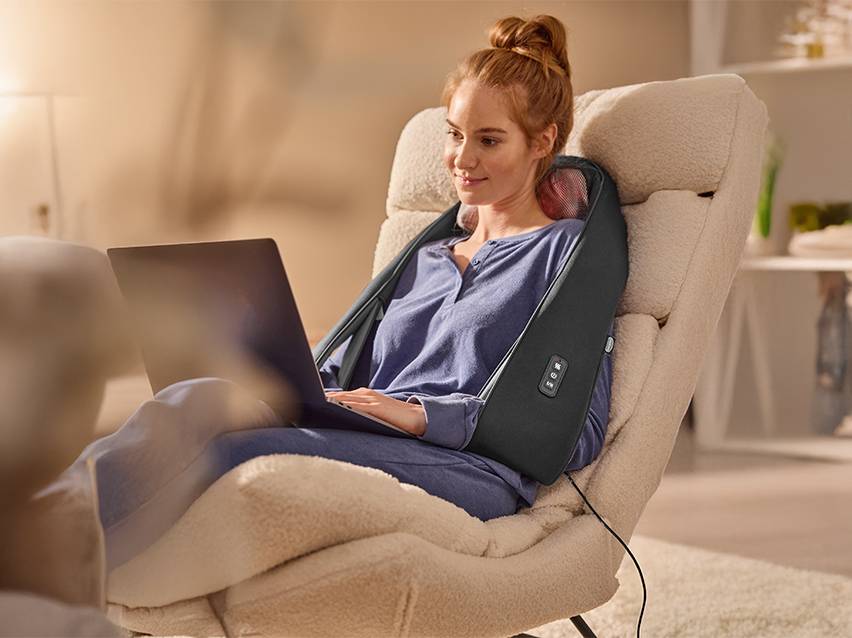 Vrouw gebruikt nek- en schoudermassageapparaat terwijl ze op een laptop werkt.