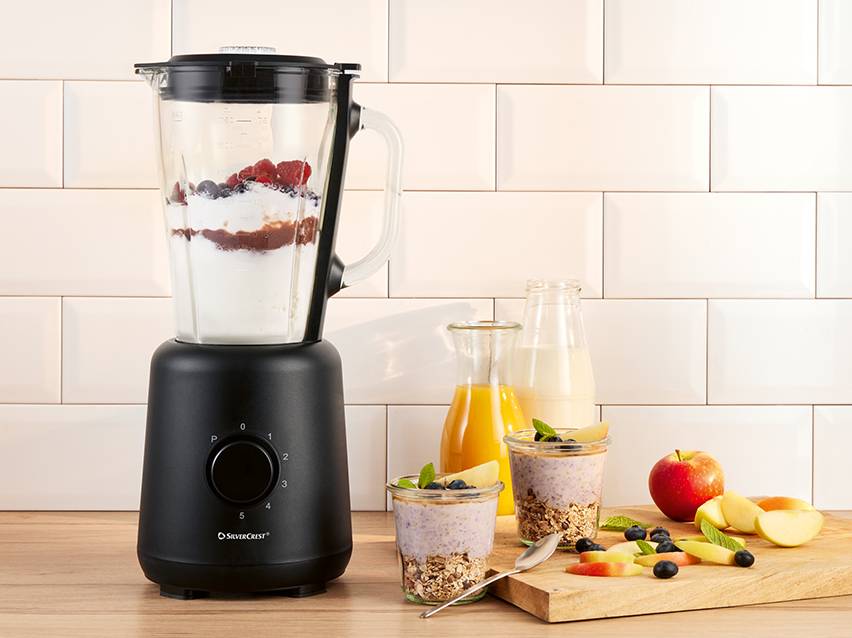 Zwarte blender met fruit en yoghurt, klaar voor een smoothie op een houten aanrecht.