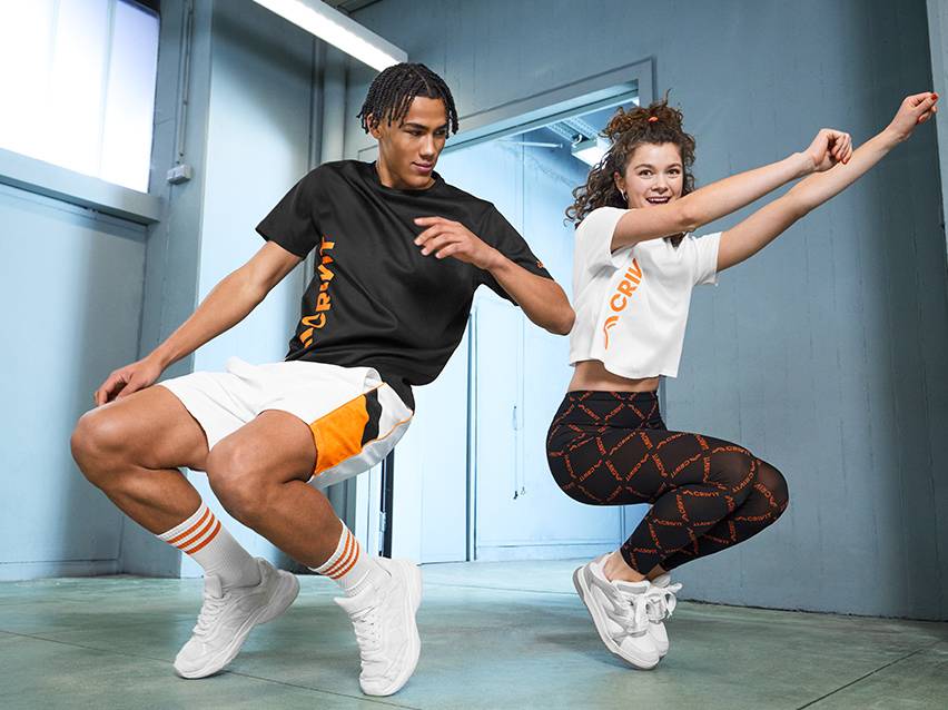 Twee mensen in sportkleding met oranje accenten, de man draagt een zwart T-shirt en witte shorts, de vrouw een wit crop top en zwarte leggings.
