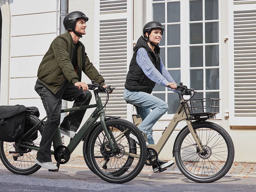 Twee mensen fietsen op elektrische fietsen, de man heeft een fietstas en de vrouw een mand.