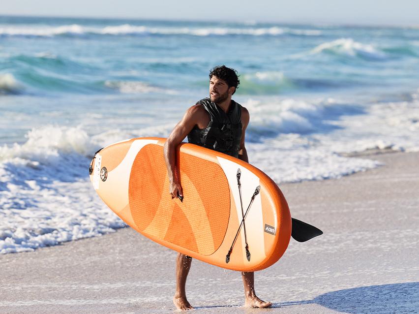 Man met zwemvest draagt een oranje paddleboard op het strand.