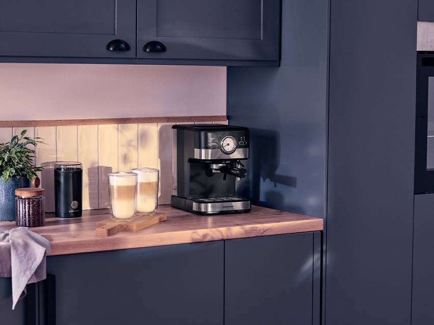 Espressomachine met twee glazen latte macchiato, koffiemolen en plant op een houten aanrecht.