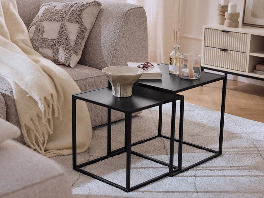 Twee zwarte bijzettafels met decoraties, een grijze bank met een plaid en een kussen.