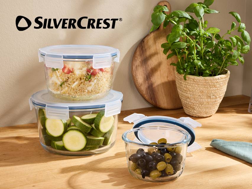 Glazen bewaarbakjes met deksels, gevuld met couscous, courgette en olijven, op een houten aanrecht.