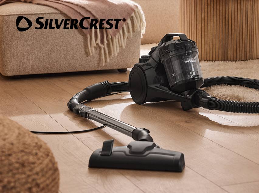 Zwarte zakloze stofzuiger op een houten vloer, met een poef en tapijt op de achtergrond.