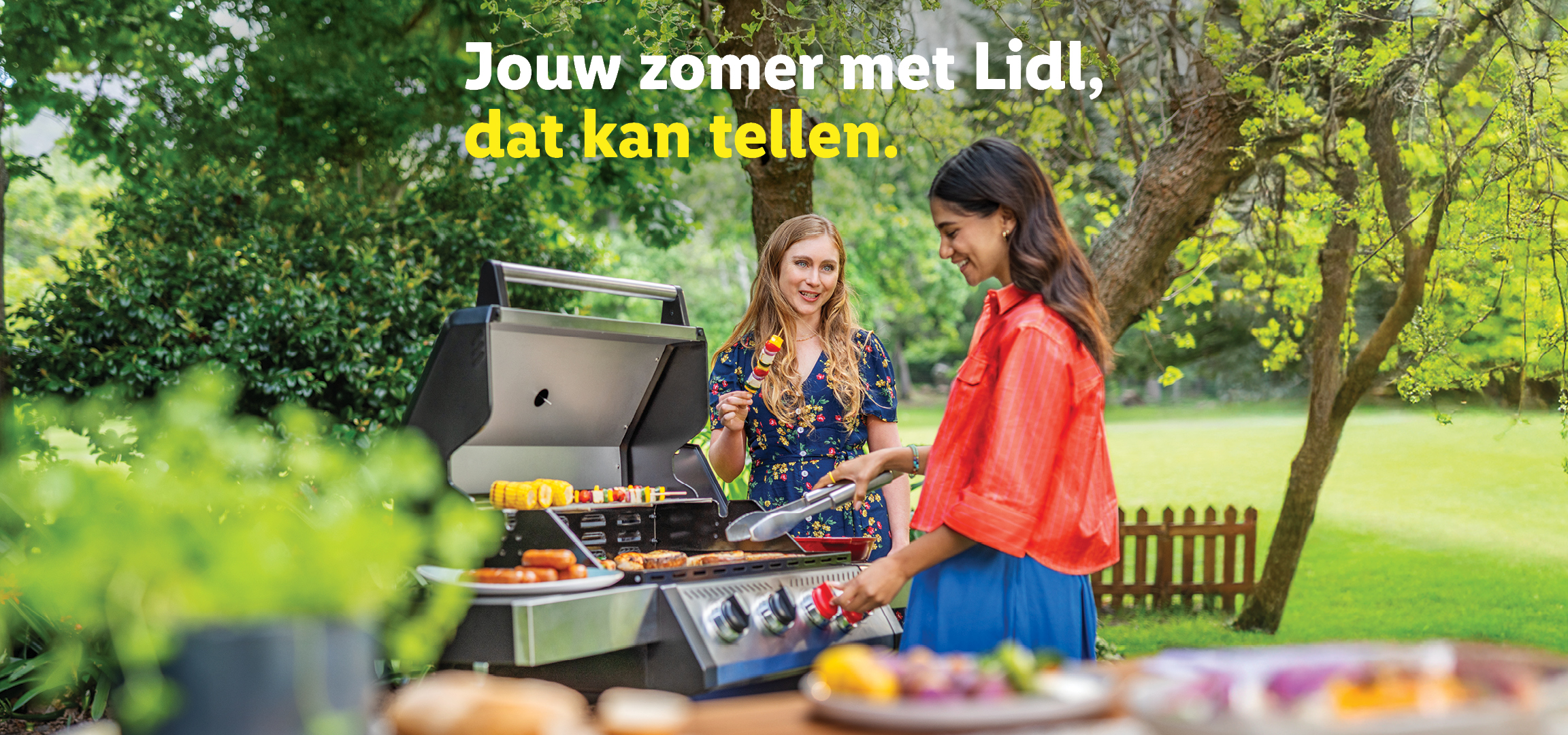 Twee vrouwen barbecueën in de tuin met groentespiesen, maïskolven en worstjes. Tekst: Jouw zomer met Lidl, dat kan tellen.