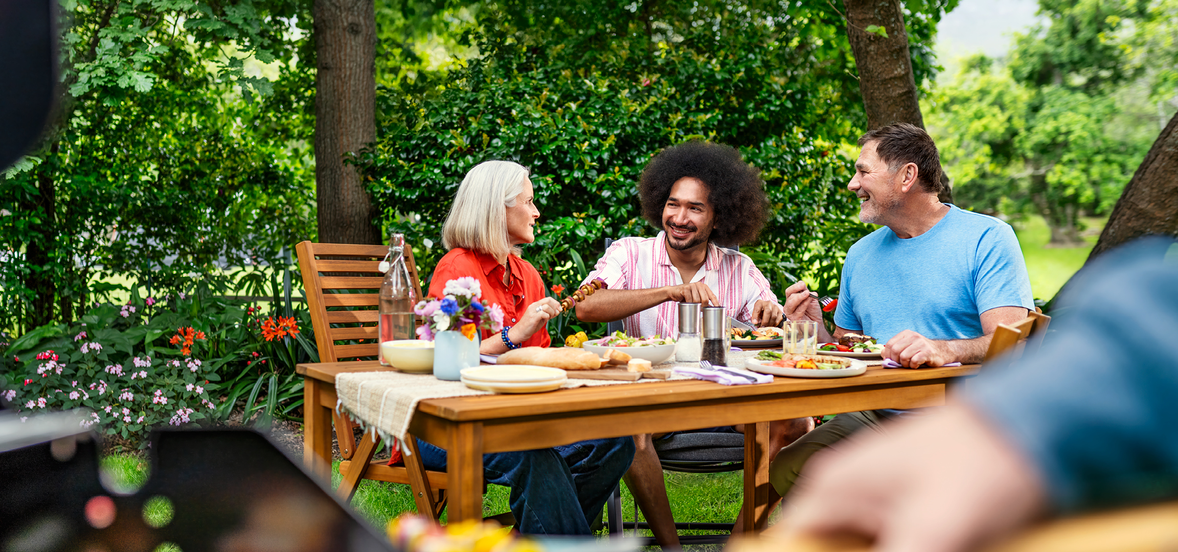 Drie mensen genieten van een maaltijd met salade, brood en spiesjes aan een houten tafel in een tuin.