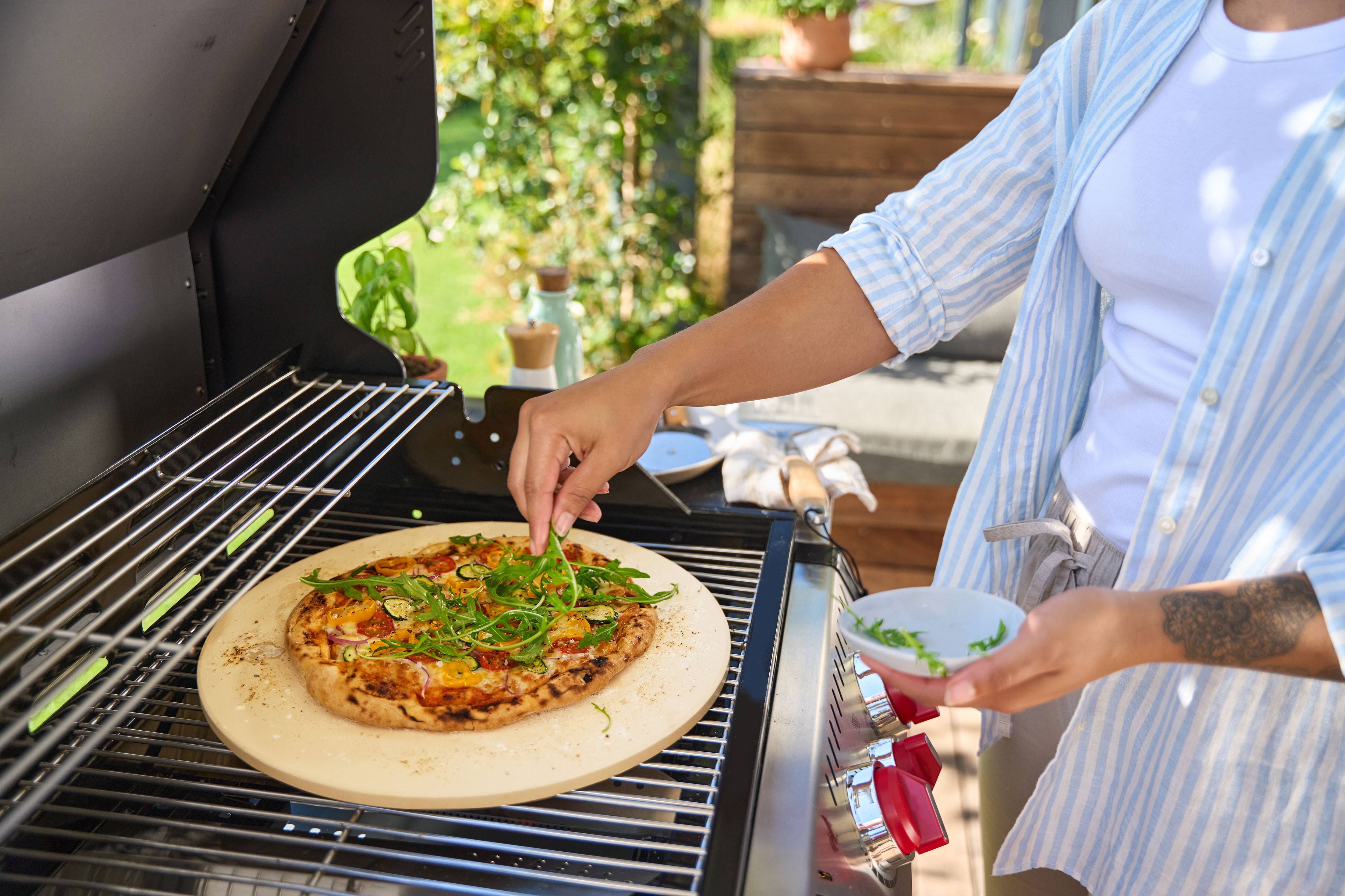 Persoon voegt rucola toe aan pizza op pizzasteen in gasbarbecue.
