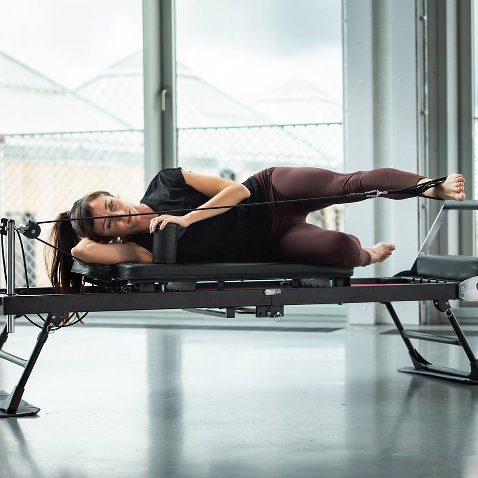 Vrouw doet Pilates op een reformer, liggend op haar zij met beenbanden.