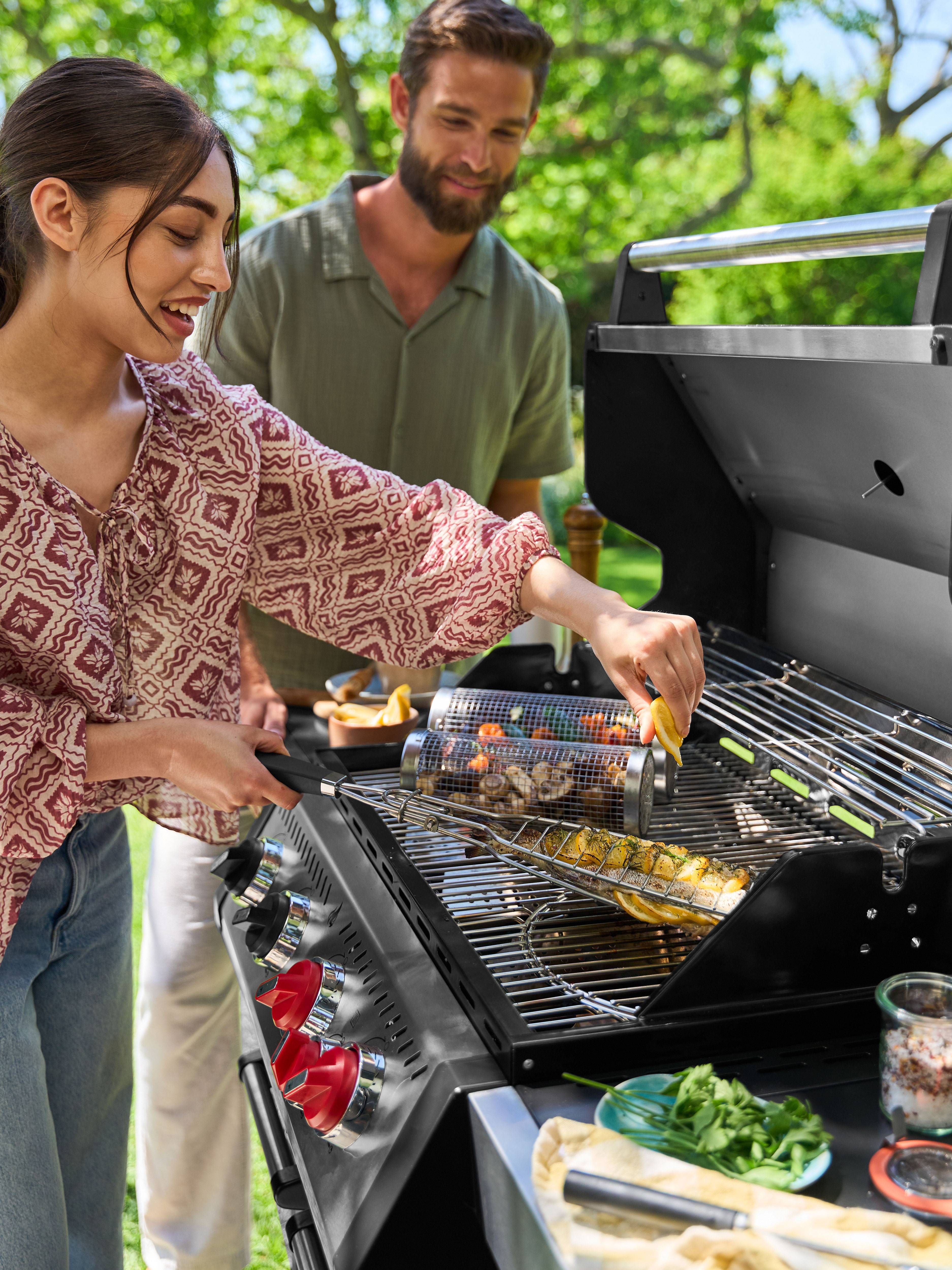 Lachende vrouw grilt vis en groenten op gasbarbecue, man kijkt van achteren toe.