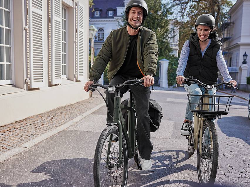 Twee lachende mensen op elektrische fietsen met helmen, op een geplaveide straat.