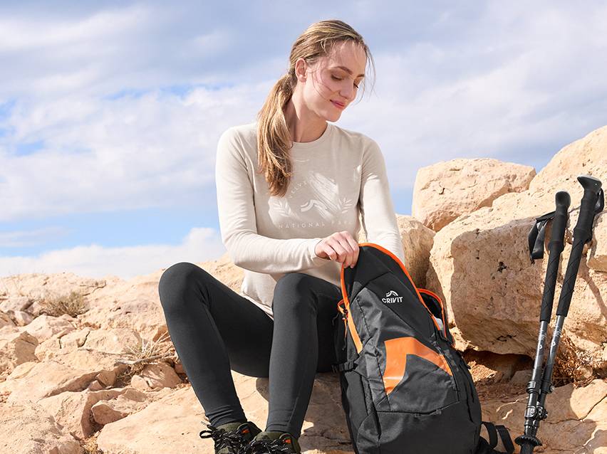 Vrouw zit op rotsen, opent een zwarte en oranje wandelrugzak, met wandelstokken ernaast.