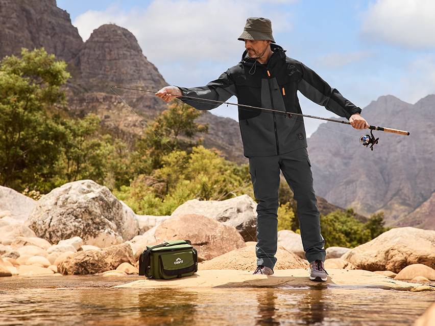 Een visser in viskleding, met een jas en broek, aan de oever van een rivier.