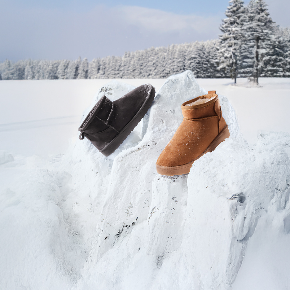 Twee warme winterlaarzen, één bruin en één donkergrijs, staan op besneeuwde rotsen in een winterlandschap.