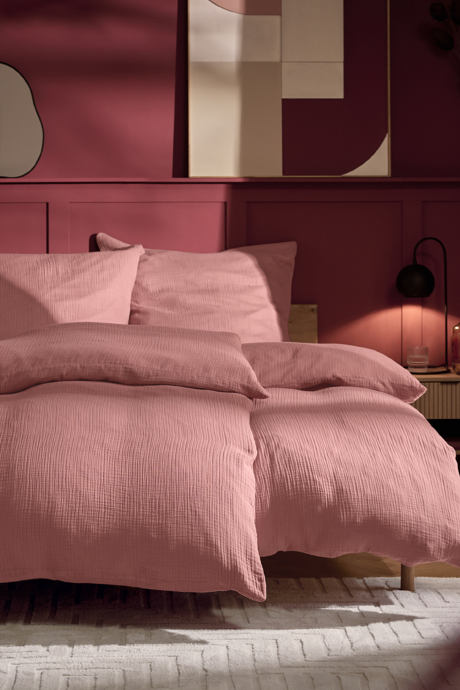 Een slaapkamer met een bed opgemaakt met roze beddengoed, tegen een bordeauxrode muur.
