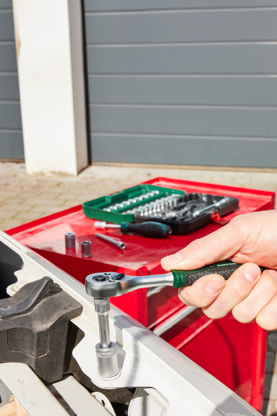 Een hand gebruikt een ratelsleutel met een dopsleutel om aan een auto te werken, met een gereedschapskist op de achtergrond.