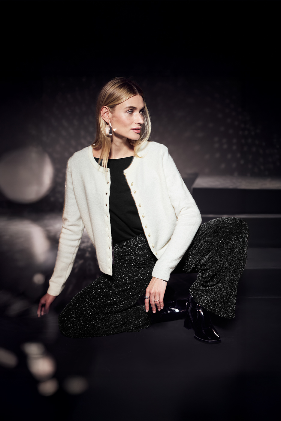 Vrouw in een wit vest met parelknoopjes, zwarte top en glimmende broek, zittend.