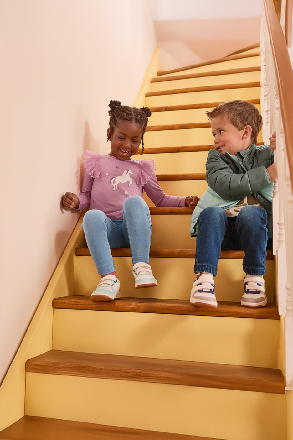 Twee lachende kinderen, gekleed in casual kleding en sneakers, zittend op een trap.