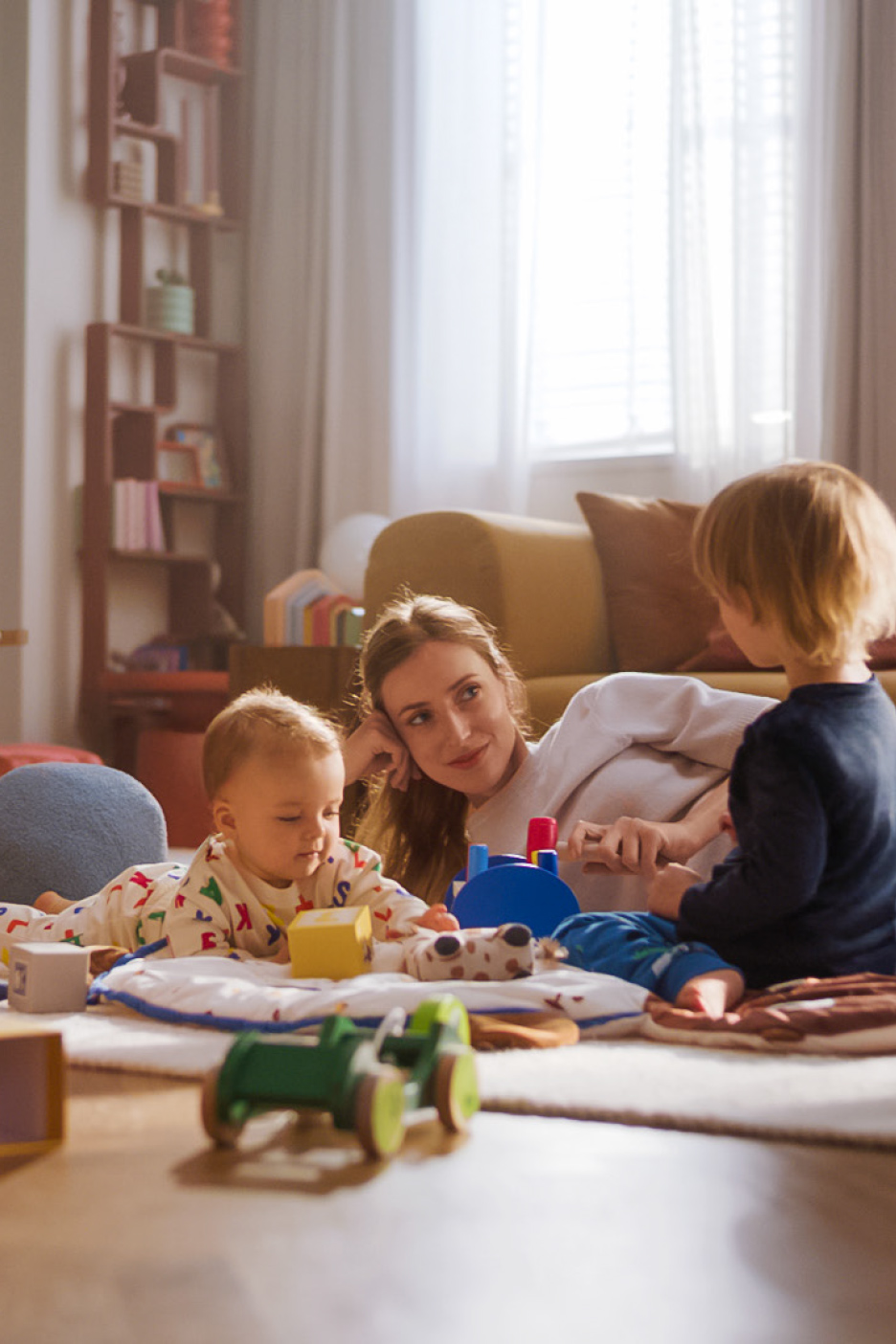 Moeder en twee kinderen spelen met speelgoed op een kleed in de woonkamer.