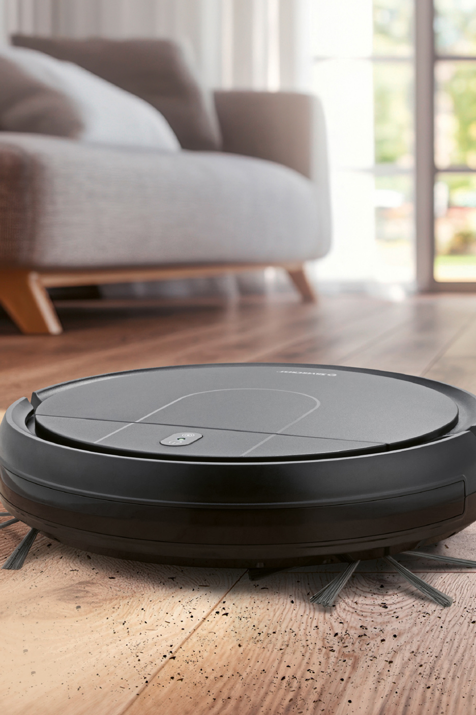 Zwarte robotstofzuiger op een vuile houten vloer, met een bank op de achtergrond.